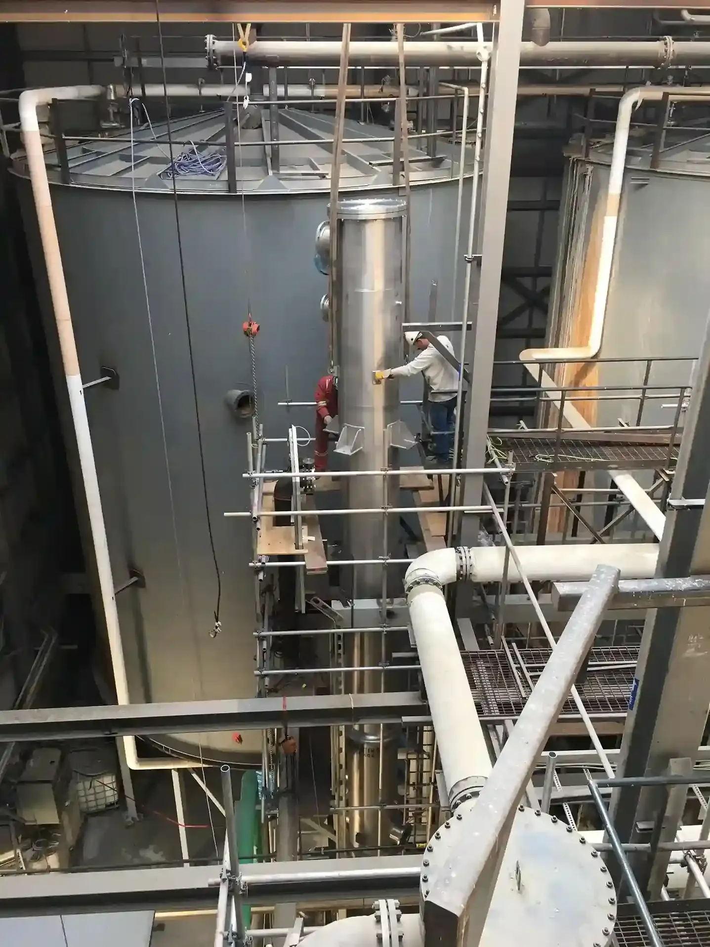 Workers on scaffolding inside an industrial plant with large tanks and metal pipes.