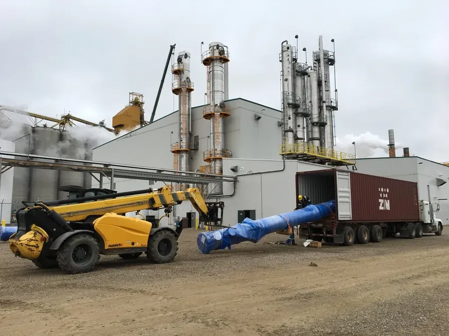 A forklift-like machine carying a pipe out of a truck in an industrial setting in front of a factory.